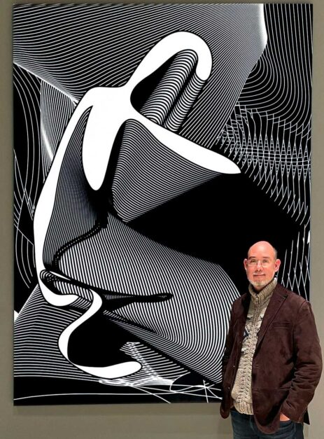 This is a photograph of artist Colin Goldberg in Hearst Tower, NYC, with his work Kneeling Icon. Kneeling Icon is an example from the artist's Metagraph Series, a body of vector-based digital drawings begun in 1999. The piece was digitally drawn in 2004 and produced as a large-scale physical work in 2022. Kneeling Icon has an AR (augmented reality) component, in which the physical work animates when viewed through an AR app on a mobile device, transforming into a 23-second-long animation that was minted as an NFT in 2021. Both the physical work and the NFT were acquired by Hearst as part of their corporate collection in 2022.
